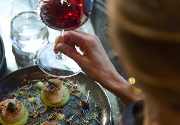 Eine Person genießt ein Glas Rotwein über einem kunstvoll angerichteten Gourmetgericht auf dunklem Teller.
