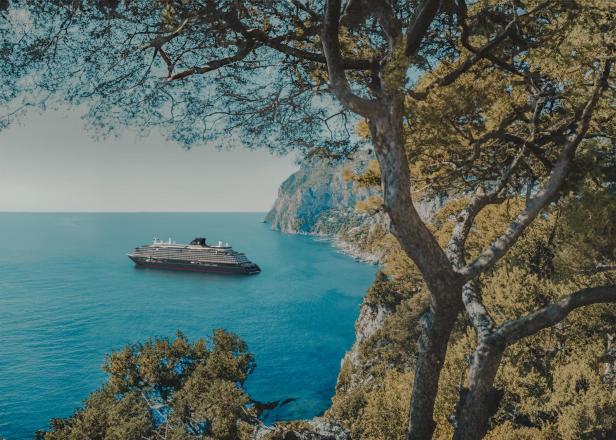 Kreuzfahrtschiff vor einer felsigen Küste, umgeben von blauem Meer und grüner Vegetation.