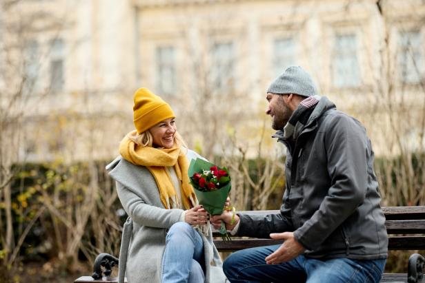 Auf einer Parkbank überreicht ein Mann einer lächelnden Frau einen Rosenstrauß.