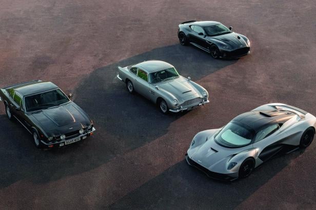 Vier verschiedene Aston Martin Sportwagen aus verschiedenen Jahrzehnten stehen auf einem asphaltierten Platz.