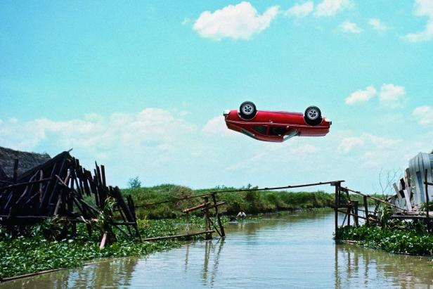 Ein rotes Auto fliegt kopfüber über einen Fluss zwischen zwei zerstörten Brückenabschnitten.