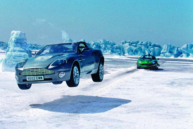 Ein silberner Aston Martin springt über eine schneebedeckte Fläche, ein grünes Auto im Hintergrund.