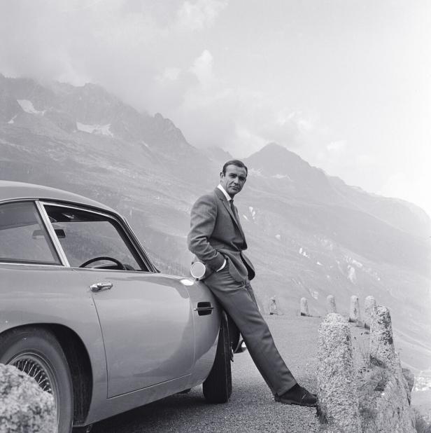 Sean Connery lehnt an einem Aston Martin vor einer Bergkulisse.