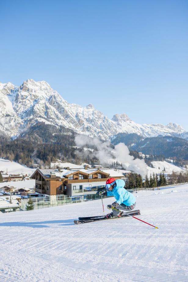 Ein Skifahrer in blauer Jacke fährt vor verschneiten Bergen und Häusern den Hang hinunter.