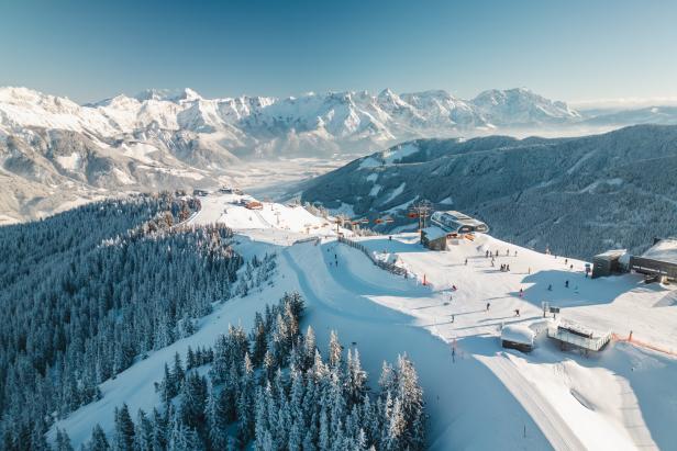 Verschneite Berge mit Skipisten, Skifahrern, Sesselliften und dichten Tannenwäldern unter blauem Himmel.
