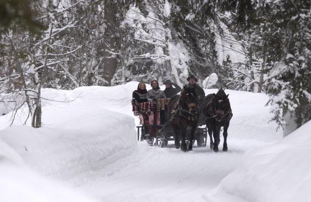 Zwei Pferde ziehen eine Kutsche mit mehreren Menschen durch einen verschneiten Waldweg.