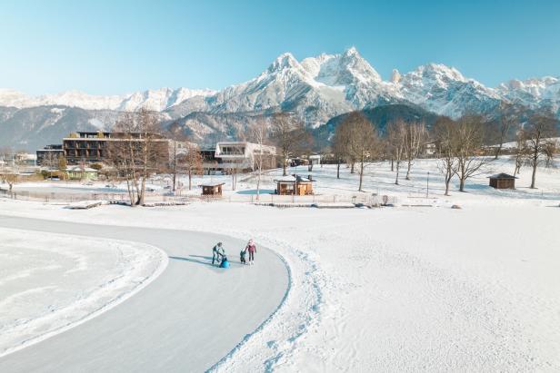 Drei Personen laufen auf einer Eisbahn im verschneiten Park, im Hintergrund schneebedeckte Berge und Gebäude.
