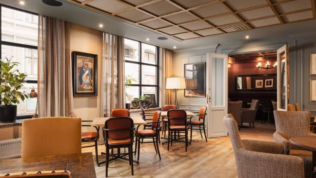 Heller, gemütlicher Raum mit Holzboden, mehreren Tischen und Stühlen, großen Fenstern, Pflanzen und stilvoller Wanddekoration.