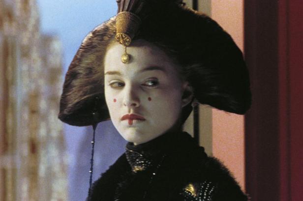 Eine Frau mit weißem Make-up, roten Punkten im Gesicht und einem kunstvollen Haarteil blickt zur Seite.