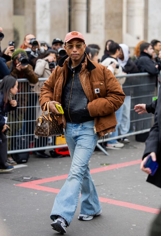Pharrell Williams trägt eine braune Jacke, Schlaghosen und eine Louis Vuitton Tasche.