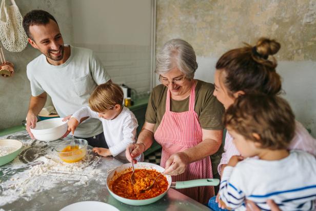 Drei Erwachsene und zwei Kinder kochen gemeinsam in einer Küche und lachen dabei.