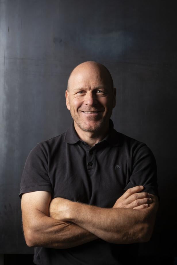 Ein lächelnder Mann mit Glatze und schwarzem Polohemd steht mit verschränkten Armen vor dunklem Hintergrund.
