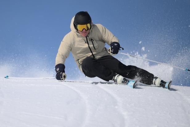 Ein Skifahrer mit Helm und Skibrille fährt auf einer schneebedeckten Piste vor blauem Himmel.