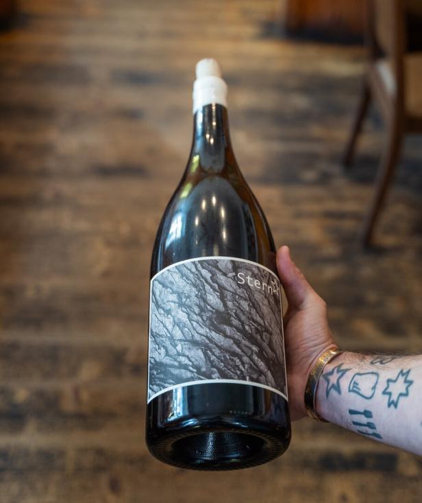 Ein tätowierter Arm hält eine große Weinflasche mit grauem Etikett und abstraktem Muster.