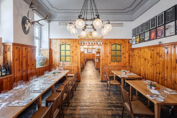Holzvertäfelter Gastraum mit langen, gedeckten Tischen, Kronleuchter und Geweih an der Wand.