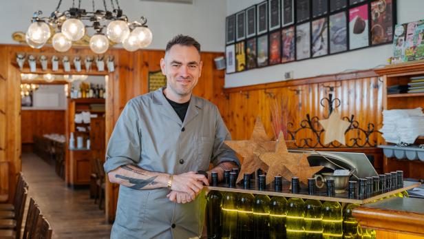 Ein Mann steht lächelnd hinter einer Theke mit Weinflaschen und Holzsternen in einem gemütlichen Restaurant.