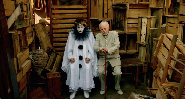 Ein Mann mit Stock sitzt neben einem Clown mit weißem Gewand vor einem Hintergrund aus Holzkisten.