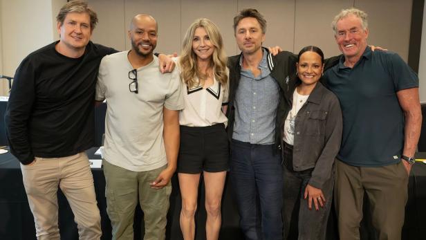 Sechs Personen stehen nebeneinander und lächeln in einem Innenraum, die Arme umeinander gelegt.