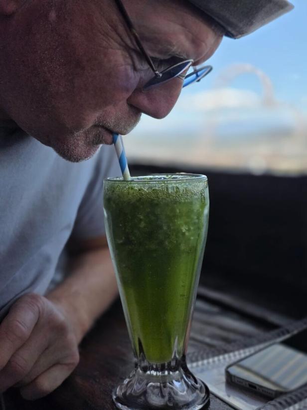 Ein Mann mit Brille trinkt durch einen Strohhalm einen grünen Smoothie aus einem hohen Glas.