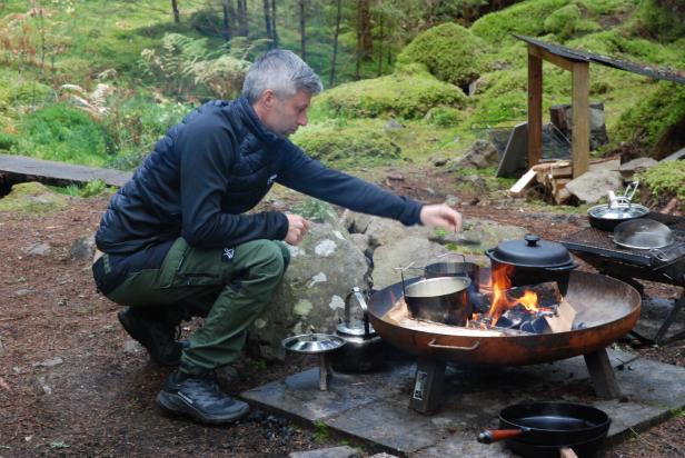 Koch Potus kocht im Wald an einer Feuerstelle mit mehreren Töpfen und Pfannen über offenem Feuer.