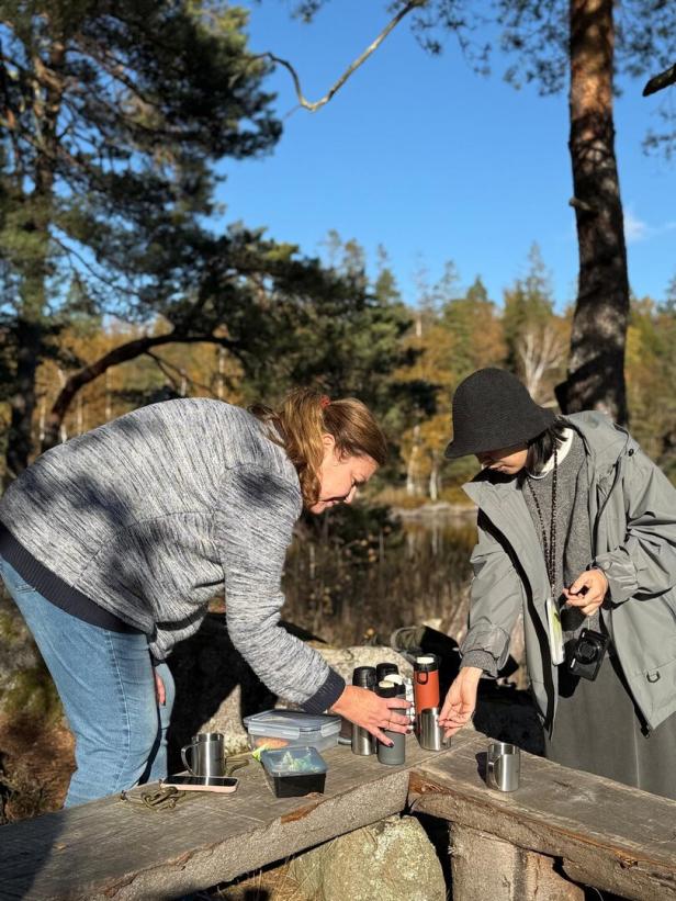 Zwei Personen schenken auf einer Holzbank im Wald Kaffee aus Thermoskannen aus.