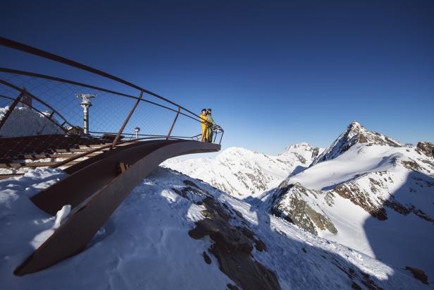 Zwei Personen stehen auf einer Aussichtsplattform mit Blick auf schneebedeckte Berge unter blauem Himmel.