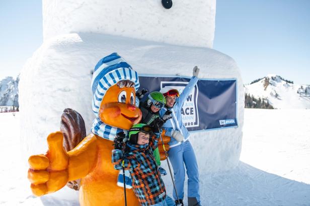 Eine Frau und zwei Kinder posieren mit einer orangefarbenen Figur vor einer Schneeskulptur im Skigebiet.