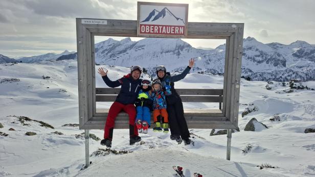 Eine Familie sitzt vor verschneiten Bergen in einem Holzrahmen mit der Aufschrift "Obertauern".