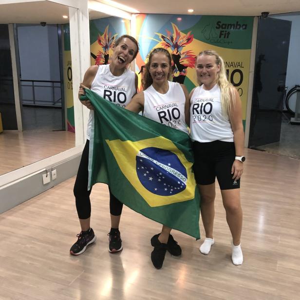 Drei Frauen in Sportkleidung halten eine brasilianische Flagge und lächeln in einem Fitnessstudio.