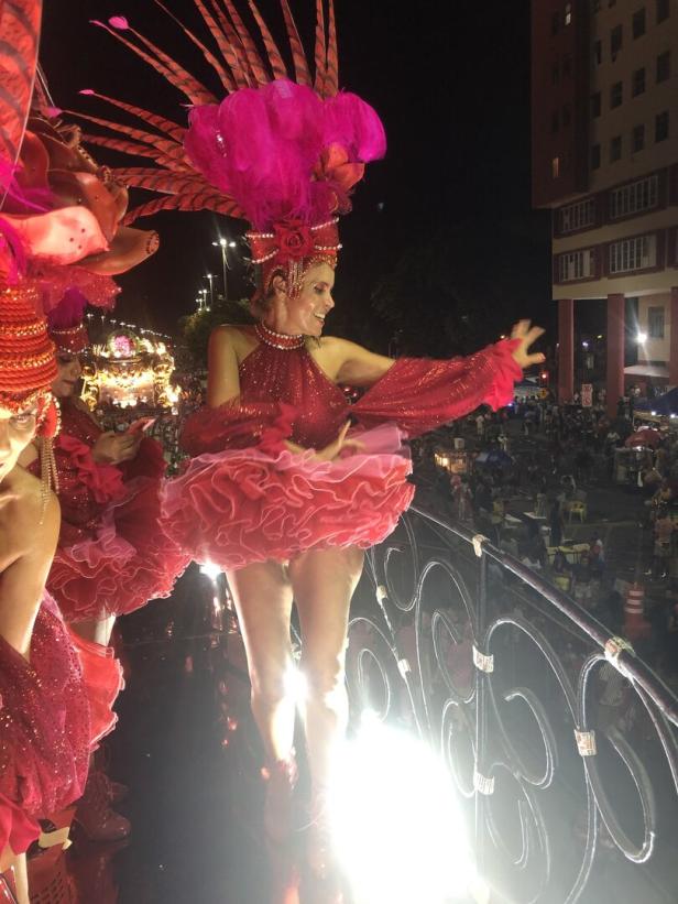 Eine Frau in rotem Karnevalskostüm mit Federschmuck tanzt auf einem Wagen vor Publikum.