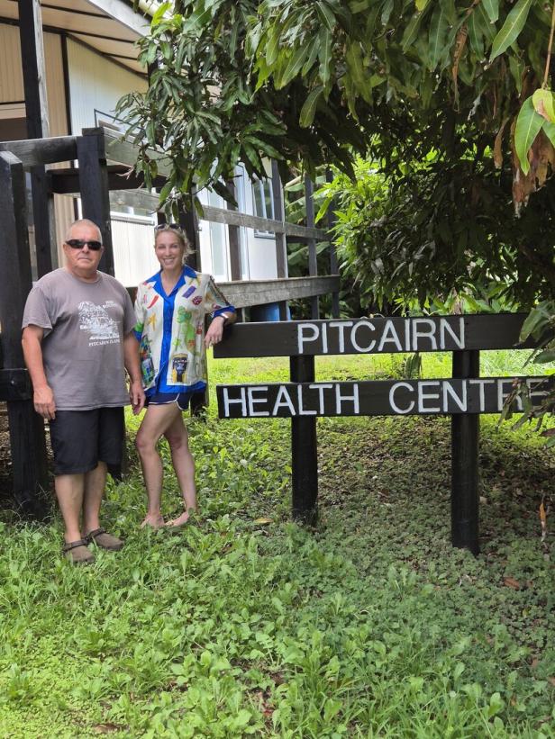 Zwei Personen stehen neben einem Schild mit der Aufschrift „Pitcairn Health Centre“ vor einem Gebäude im Grünen.