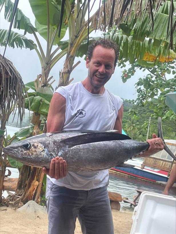 Ein Mann hält einen großen Fisch an einem tropischen Strand mit Palmen und Booten im Hintergrund.