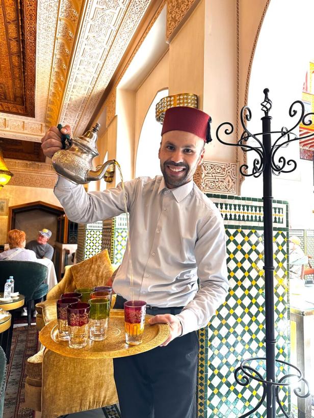 Ein lächelnder Mann mit rotem Hut gießt Tee aus einer Kanne in Gläser auf einem goldenen Tablett.