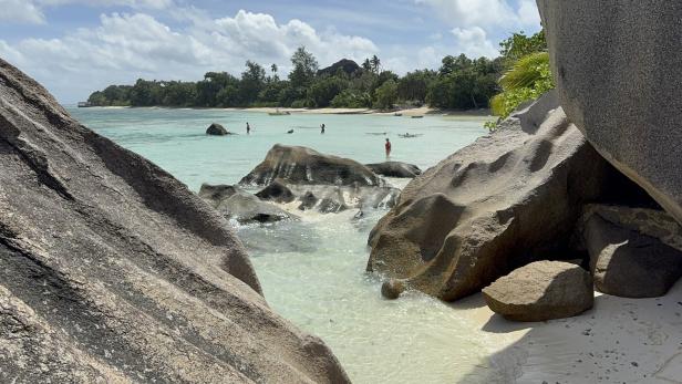 Große Granitfelsen am Rand eines türkisfarbenen Meeres, im Hintergrund Menschen im flachen Wasser und dichter grüner Wald.