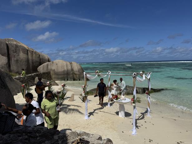 Ein Hochzeitspaar steht unter einem mit weißen Tüchern geschmückten Pavillon am Strand vor türkisfarbenem Meer und Felsen.