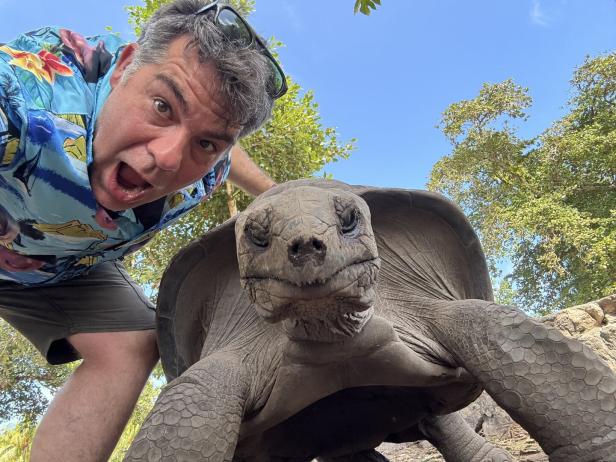 Ein Mann mit bunter Hemd macht ein Selfie mit einer großen Schildkröte im Freien unter blauem Himmel.