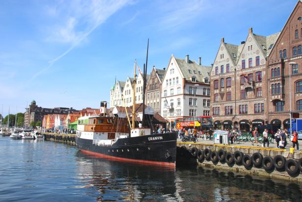Ein Schiff liegt an einer belebten Uferpromenade mit bunten historischen Häusern und vielen Menschen im Sonnenschein.