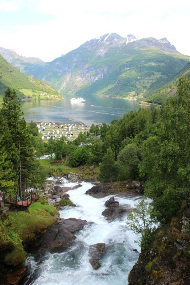 Ein Fluss stürzt durch eine grüne Landschaft, im Hintergrund ein Fjord mit einem Kreuzfahrtschiff.