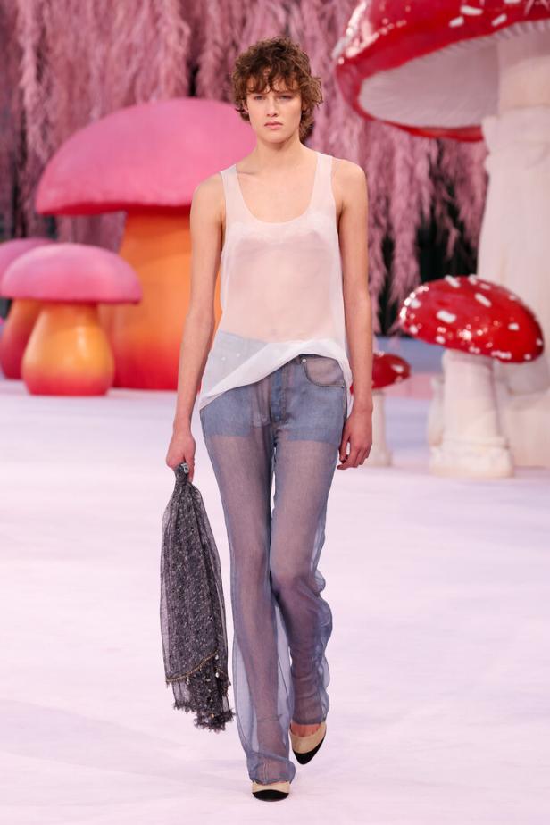 Ein Model mit lockigen Haaren trägt ein transparentes weißes Top und durchsichtige Jeans, am Laufsteg umgeben von großen, bunten Pilzen.