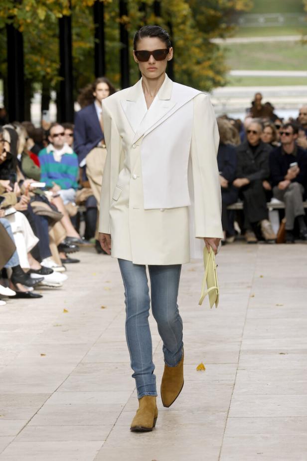 Eine Frau mit Sonnenbrille präsentiert Mode mit Blazer, skinny Jeans und Stiefeln auf einem Laufsteg.