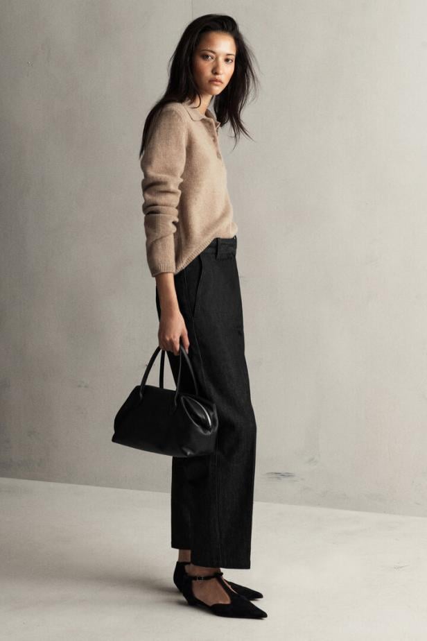Ein Model mit dunklen Haaren trägt einen beigen Pullover, dunkle Hose aus Jeans-Stoff und hält eine schwarze Handtasche.