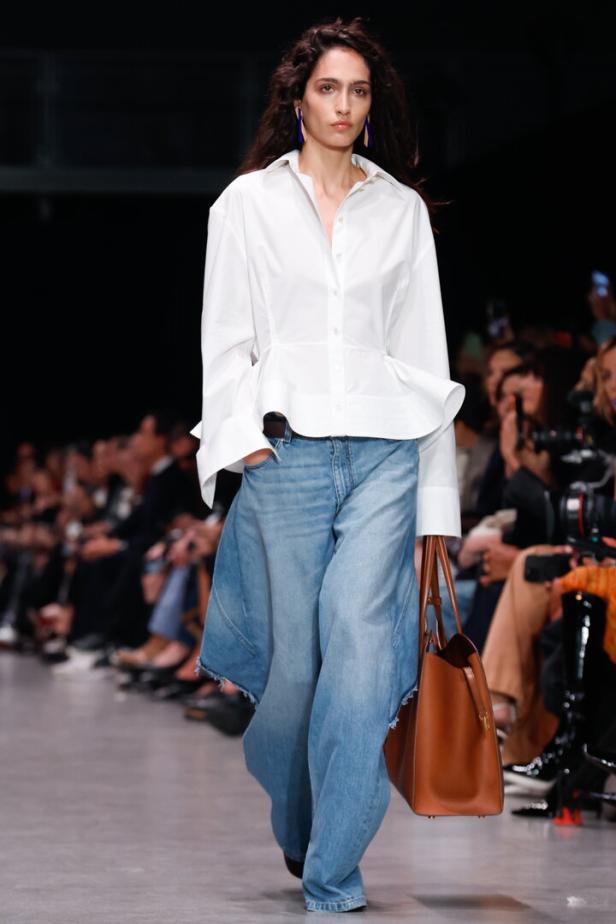 Frau mit weißer Bluse und weiter Jeans läuft mit brauner Tasche über einen Laufsteg.