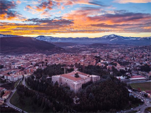 Eine große Festung ist von Bäumen umgeben, dahinter erstreckt sich eine Stadt vor schneebedeckten Bergen im Sonnenuntergang.