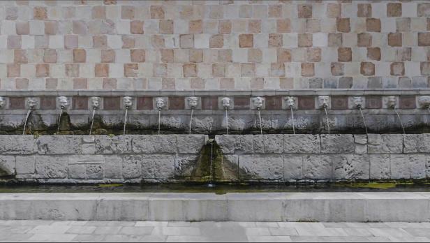 Steinerner Brunnen mit mehreren Wasserspeiern in Form von Gesichtern vor einer gemusterten Wand.