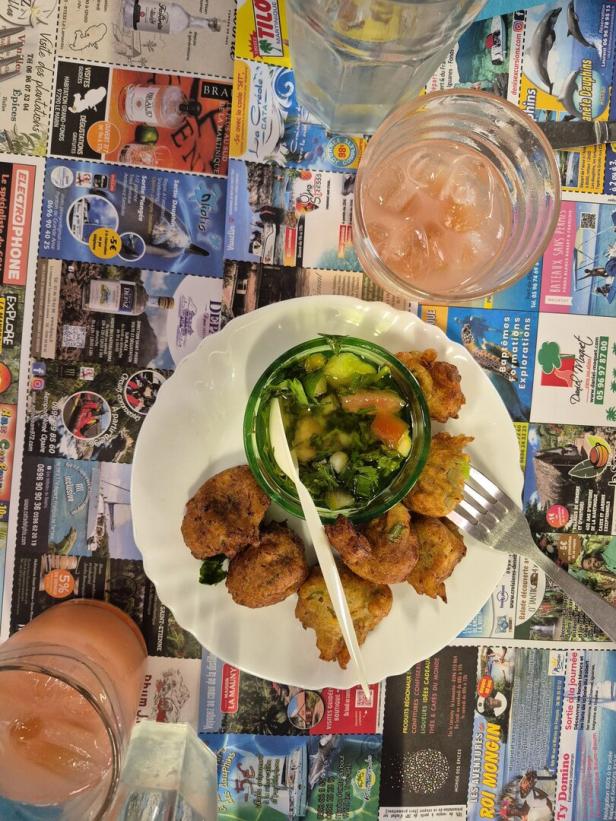 Ein Teller mit frittierten Bällchen und einem Glas mit Gemüse, dazu zwei Gläser mit rosa Getränk und Eiswürfeln auf einem bunten Untergrund.