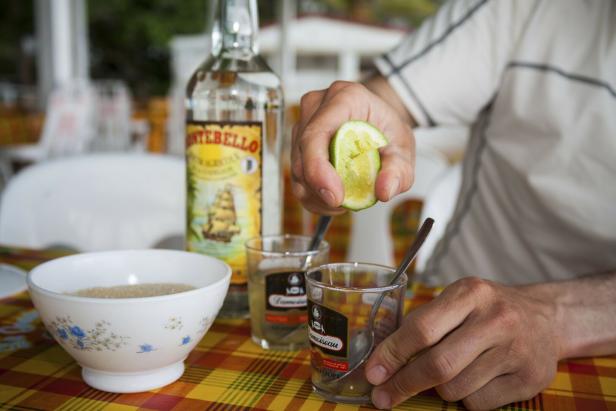 Eine Person presst Limette in ein Glas mit Rum, daneben stehen eine Flasche Rum und eine Schale mit Zucker auf einem bunt karierten Tisch.