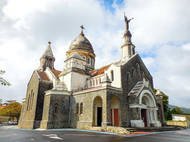 Steinkirche mit Kuppel, Türmen und Statuen, umgeben von einem leeren Parkplatz und bewölktem Himmel.