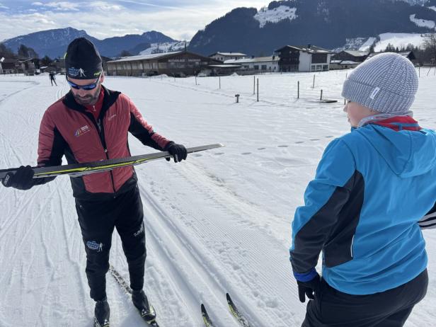 Ein Erwachsener zeigt einem Kind auf einer verschneiten Loipe einen Langlaufski, Berge und Häuser im Hintergrund.