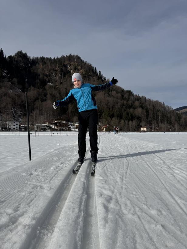Ein Kind fährt auf Langlaufskiern durch eine verschneite Landschaft mit Bergen und Bäumen im Hintergrund.
