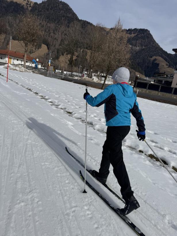 Eine Person in blauer Jacke und Skiern gleitet über eine verschneite Fläche vor einer Bergkulisse.